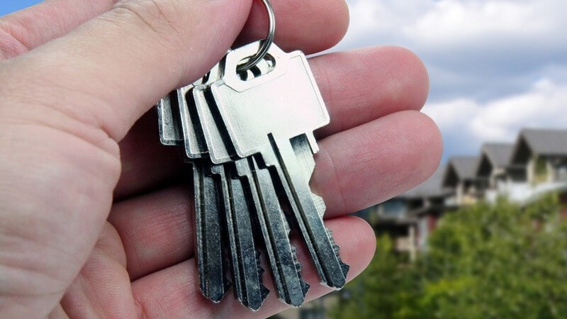 Linke Hand eines Mannes hält Haustürschlüssel; Himmel, Bäume und mehrere Einfamilienhäuser im Hintergrund