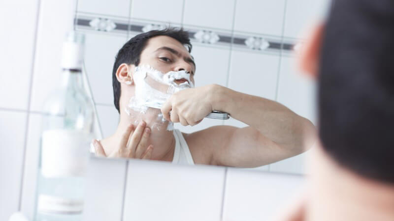 Blick durch Spiegel: Dunkelhaariger Mann rasiert sich den Bart mit Rasiermesser (Nassrasur)