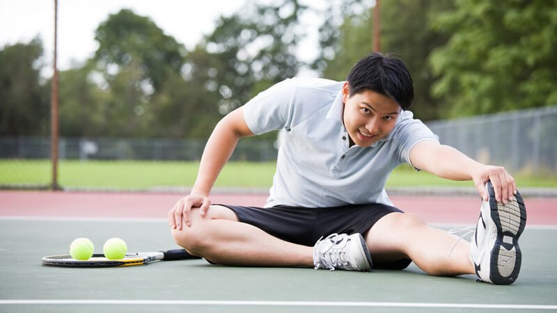Junger Mann im Sportoutfit sitzt auf Tennisplatz und dehnt sich vor dem Tennisspiel