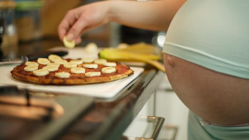 Schwangere Frau belegt Pizza mit Bananenscheiben