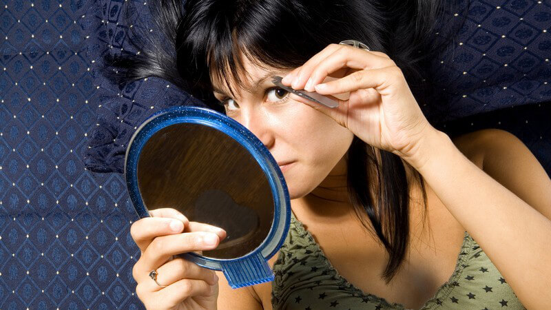 Junge, dunkelhaarige Frau zupft Augenbrauen vor Kosmetikspiegel