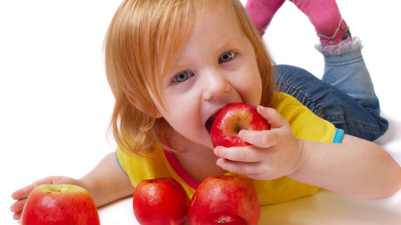 Kleines rothaariges Mädchen liegt auf dem Bauch und beisst in roten Apfel rein
