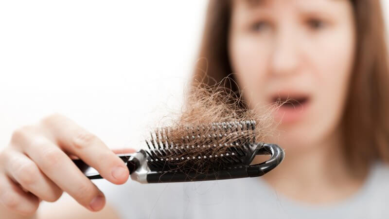 Frau mit entsetztem Blick hält Haarbürste mit Haarbüscheln daran, Haarausfall