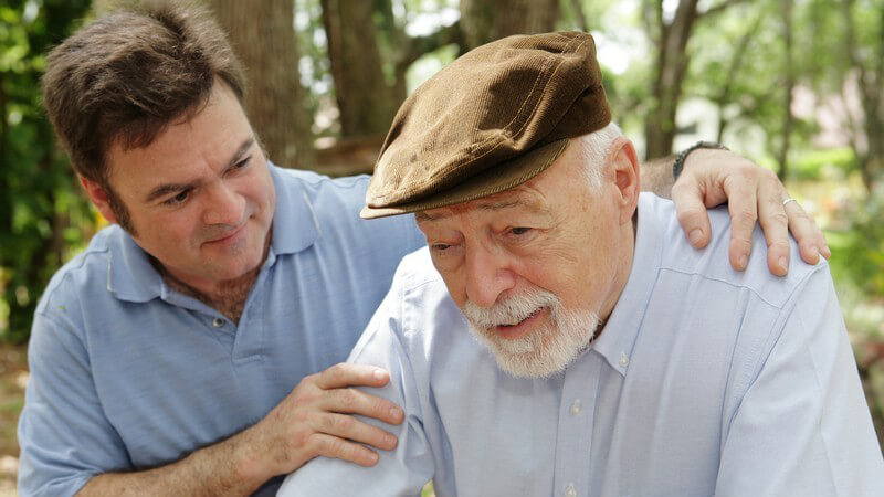 Alter Mann mit braunem Hut, der von jungem Mann an den Schultern gestützt wird im Park