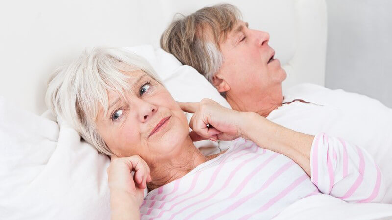 Älteres Ehepaar liegt im Bett, der Mann schnarcht, die Frau hält sich genervt die Ohren zu