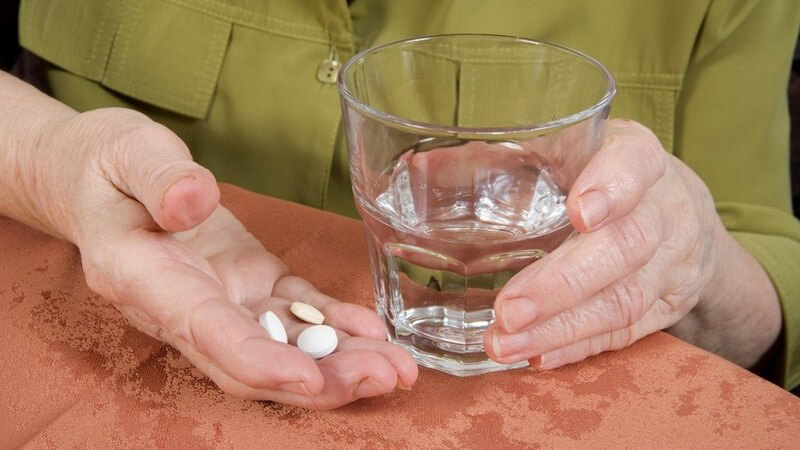 Ein Wasserglas und 3 weiße Tabletten, gehalten von alten Händen auf beiger Tischdecke vor grüner Bluse