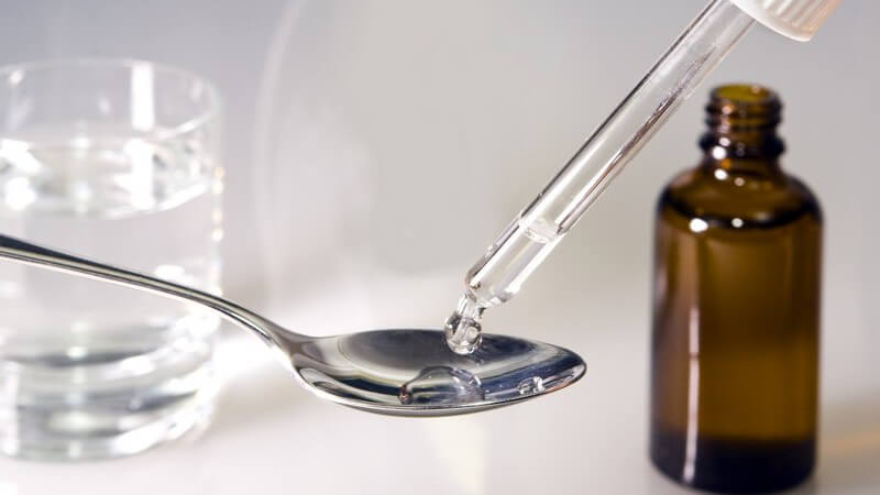 Medizin wird aus Pipette auf Löffel getropft, im Hintergrund Glas mit Wasser