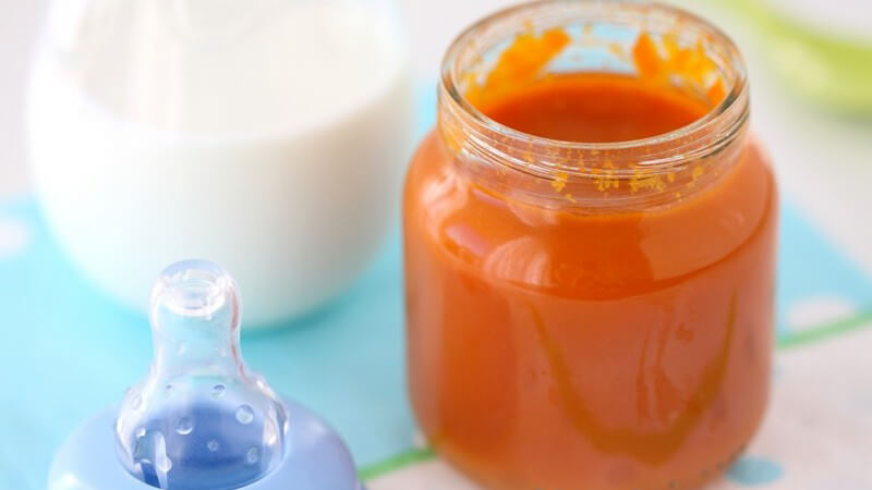 Orangener Babybrei im Glas, daneben Nuckel einer Trinkflasche auf hellblauem und weißem Boden