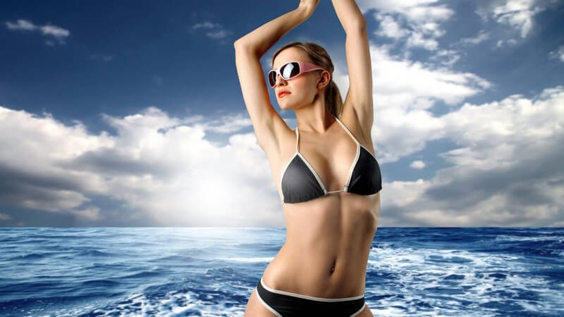 Junge Frau im schwarzen Bikini mit Sonnenbrille vor Wasser unter blauem Himmel