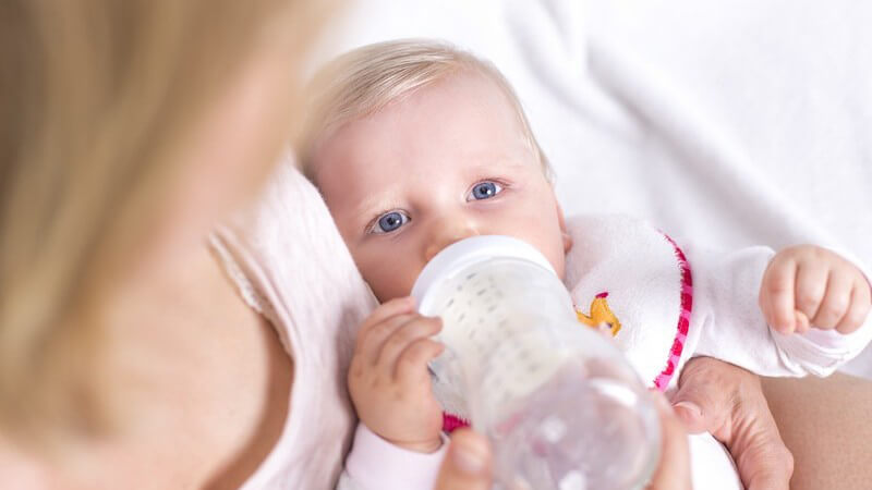Blondes Baby mit Lätzchen liegt bei der Mutter im Arm und trinkt aus einem Fläschchen