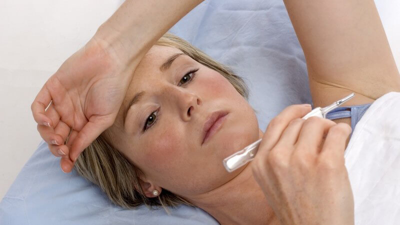 Junge Frau liegt im Bett, hat einen Arm über Stirn gelegt, hält Fieberthermometer fest
