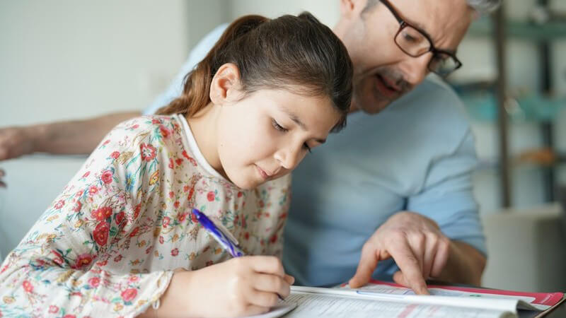 Vater mit grauen Haaren und Brille hilft seiner Tochter bei den Hausaufgaben und zeigt an eine Stelle im Schulbuch