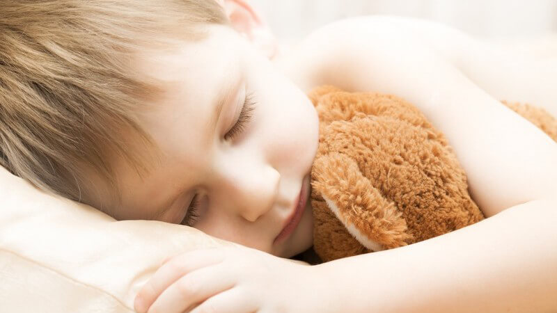 Kleiner Junge liegt mit Teddy im Arm im Bett und schläft