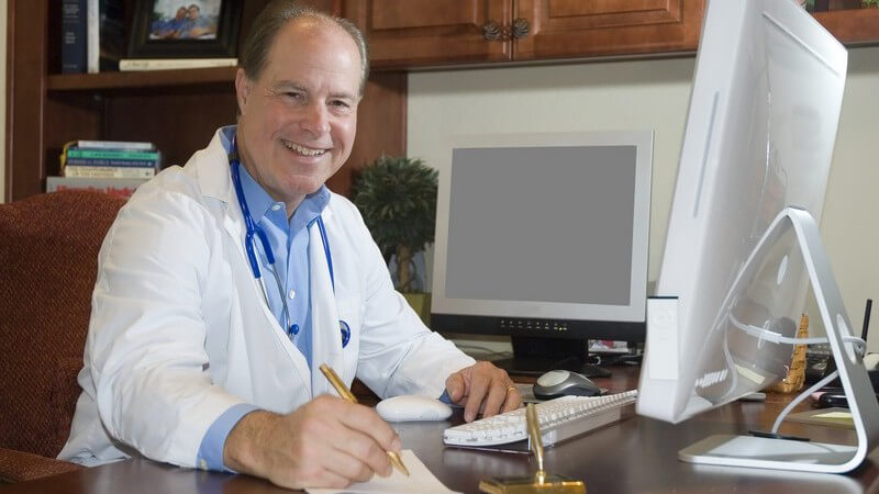 Arzt in weißem Kittel sitzt in Büro am Computer und lacht in Kamera