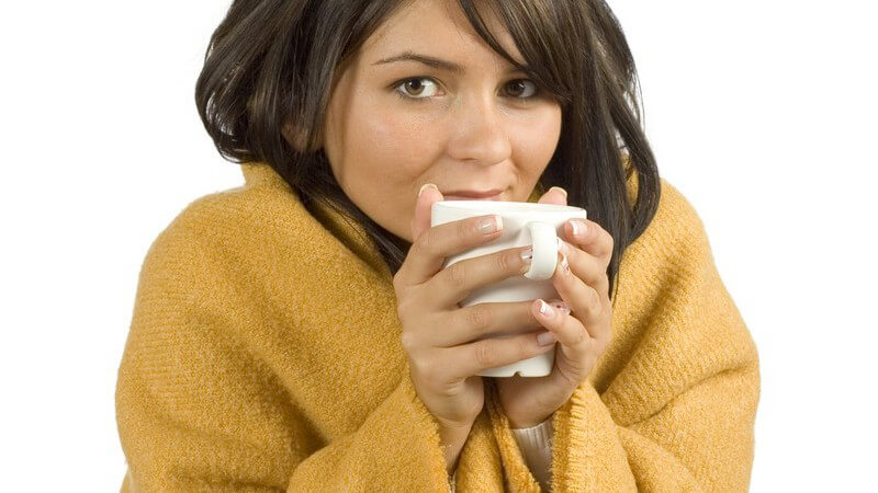 Dunkelhaarige, junge Frau mit brauner Wolldecke und weißer Tasse friert und zittert