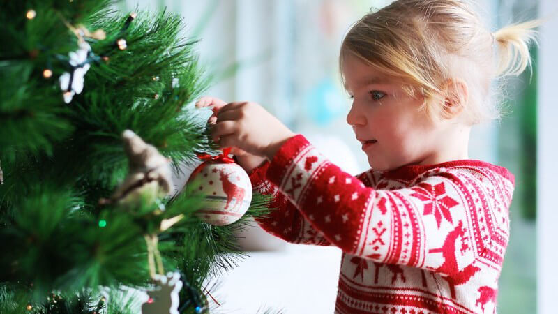 Kleines blondes Mädchen in rotem Winterpulli schmückt den Weihnachtsbaum mit Christbaumkugeln