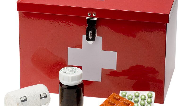 Rote Erste-Hilfe-Metallkiste (Hausapotheke) mit weißem Kreuz, davor liegen Tabletten und ein Verband