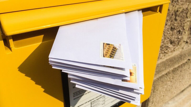 Ein Stapel Briefe steckt im Schlitz eines gelben Postkastens