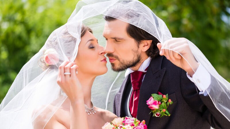 Hochzeitspaar küsst sich unter dem Brautschleier im Grünen
