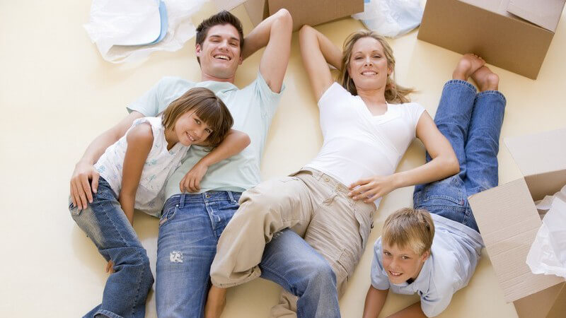 Junge lächelnde Familie liegt im neuen Zuhause zwischen Umzugskartons auf dem Boden