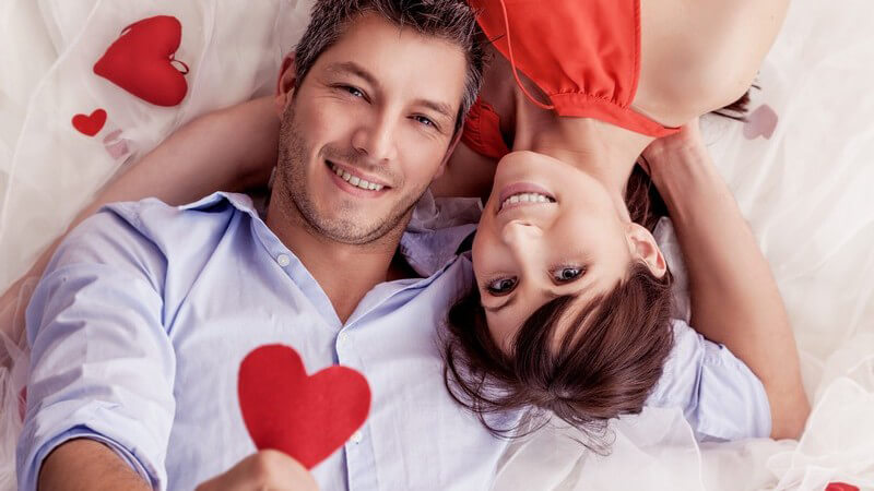 Junges Paar liegt auf dem Bett und hält ein rotes Herz hoch