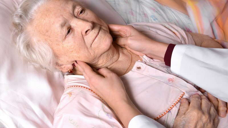 Arzt untersucht eine im Bett liegende alte Frau durch Abtasten der Lymphknoten am Hals