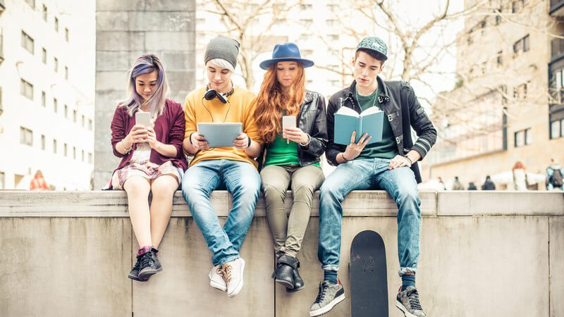 Vier Jugendliche sitzen nebeneinander auf einer Mauer und beschäftigen sich mit Smartphone, Tablet und Buch