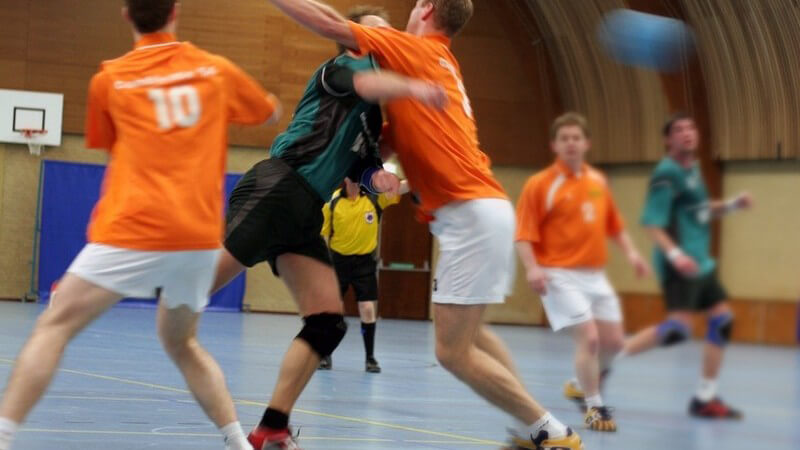 Handballspiel in Turnhalle, zwei Mannschaften, Zusammenstoß von zwei Spielern, Handball rechts im Bild