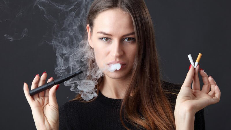 Dunkelhaarige Frau vor schwarzem Hintergrund dampft eine E-Zigarette und hält eine geknickte Filterzigarette in der Hand