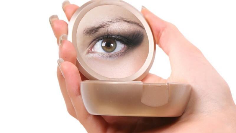 Frauenhand hält Kosmetikspiegel, im Spiegelbild ihr linkes Auge