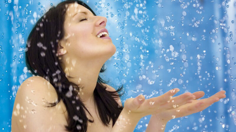 Schwarzhaarige Frau hält ihre Hände vor den Körper, um heruntertropfendes Wasser in blauer Dusche aufzufangen