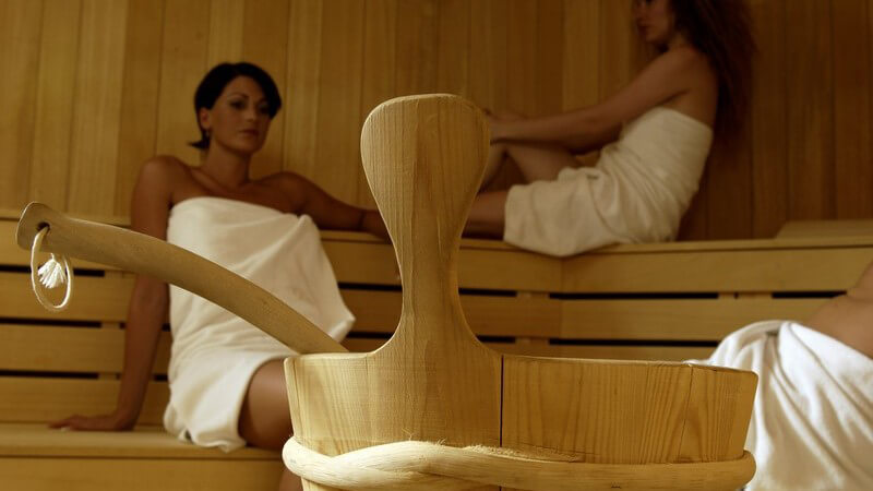 Zwei Frauen in weißen Handtüchern sitzen in Sauna, im Vordergrund Aufguss Gefäß