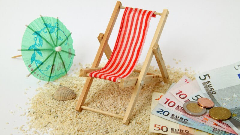 Mini-Sonnenliege auf Sand, daneben Gelscheine - Urlaubsgeld
