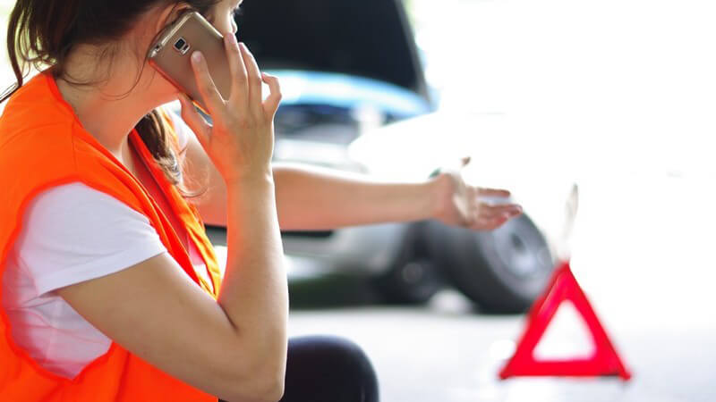 Frau in roter Warnweste telefoniert nach einem Autounfall oder einer Panne mit dem Handy, vor ihr steht ein Warndreieck