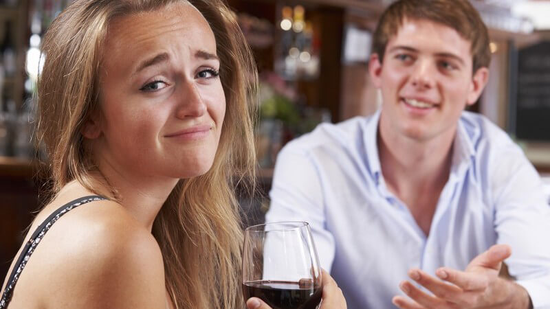 Zwei junge Leute bei einem Blind-Date, sie schaut enttäuscht und abgeneigt in die Kamera und hält ein Glas Rotwein