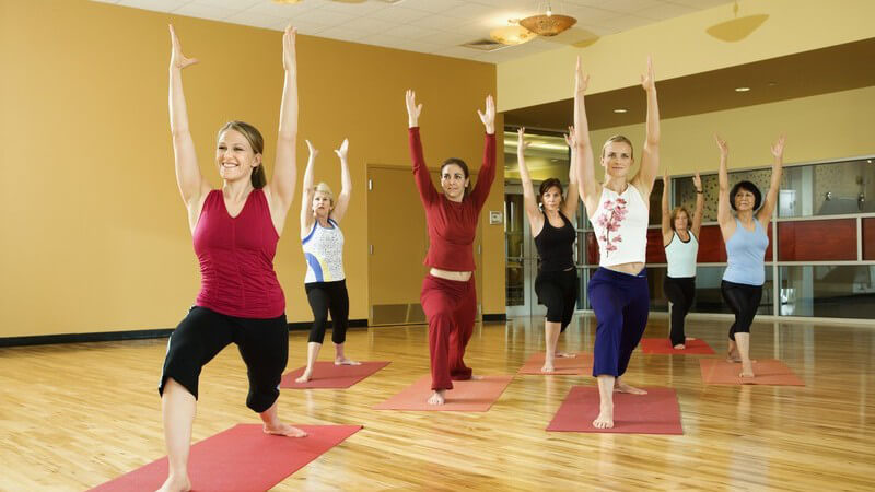 7 Frauen auf Yogamatten, die Hände nach oben, ein Bein nach vorn, eins nach Hinten stehend
