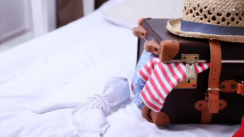 Brauner Reisekoffer liegt leicht geöffnet auf einem Bett, auf dem Koffer ein Sonnenhut