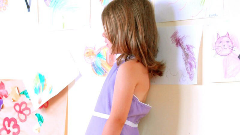 Kleines Mädchen in lila Kleid lehnt an Wand mit gemalten Kinderbildern