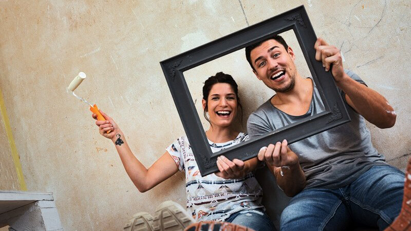 Lachendes Paar posiert vor kahler Wand in neuer Wohnung mit einem vorgehaltenen Bilderrahmen