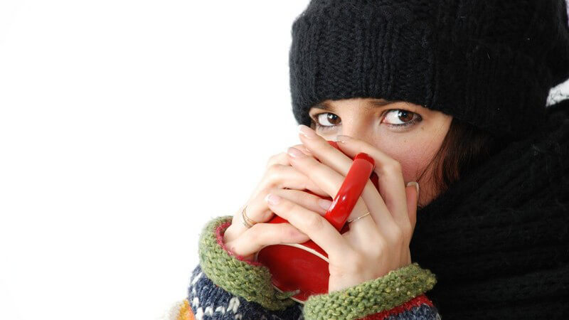 Frau in Winterkleidung mit Schal und Mütze trinkt aus Becher, wärmt sich Hände daran