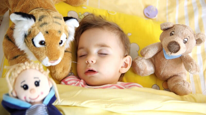 Kleinkind liegt im Bett und schläft, umgeben von Kuscheltieren