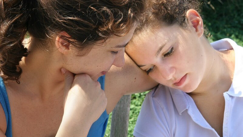 Zwei braunhaarige Freundinnen oder Schwestern sitzen draußen zusammen, wirken bedrückt und trösten sich gegenseitig