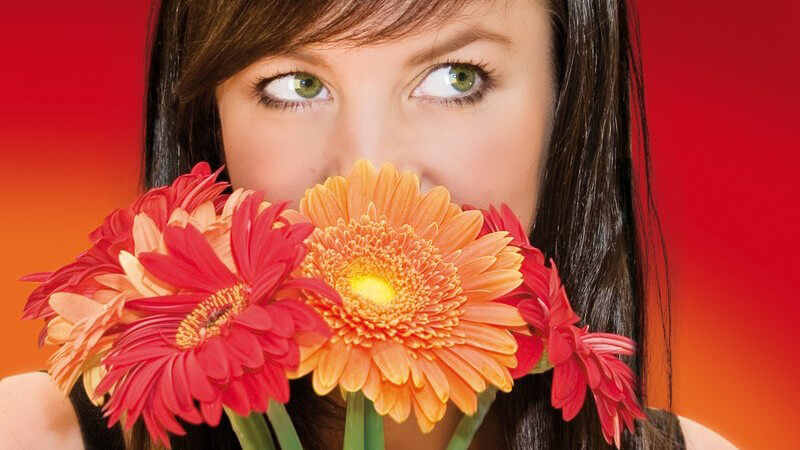 Junge, dunkelhaarige Frau versteckt ihr Gesicht halb hinter roten und orangen Blumen