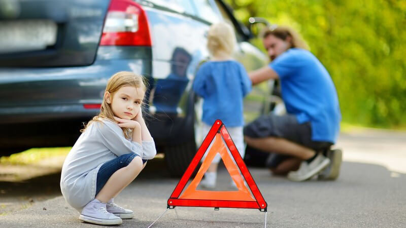 Autopanne - Mann beim Reifenwechsel auf der Straße, kleines Mädchen hockt davor neben Warndreieck