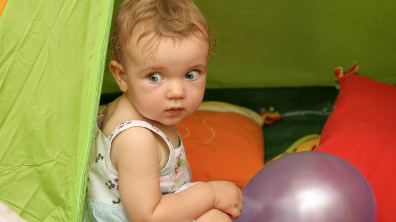 Blondes Kind in weißem Kleid sitzt in grünem Zelt mit violettem Luftballon und Kissen