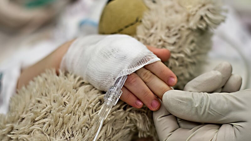 Kinderhand mit Verband und Zugang umklammert einen Teddybären im Krankenhaus
