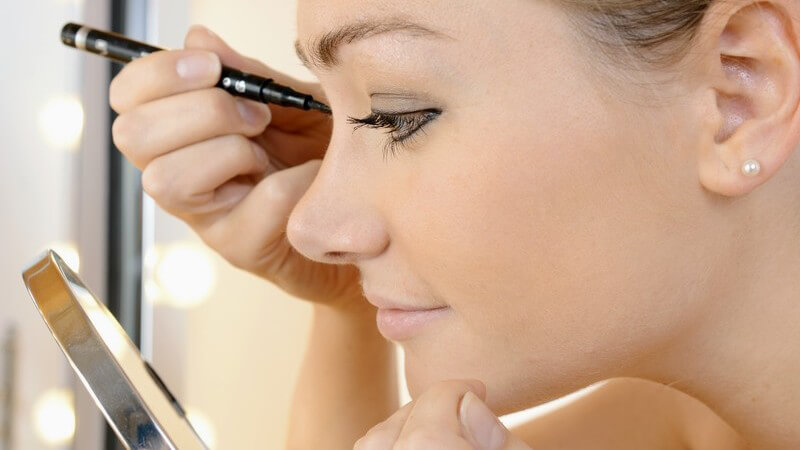 Junge Frau mit Perlenohrring schminkt ihre Augen vor einem runden Schminkspiegel