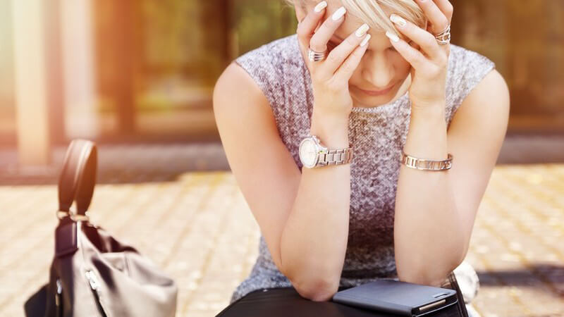 Frau mit kurzen blonden Haaren sitzt mit Handtasche, Laptop und Handy vor einem Gebäude und hält die Hände vors Gesicht