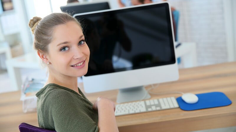 Junge Frau in olivfarbenem Shirt sitzt vor einem modernen Computer am Schreibtisch im Büro und guckt nach hinten
