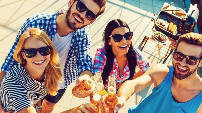 Vier Freunde in Sommerkleidung stoßen auf einer Terrasse mit Flaschenbier an, im Hintergrund steht ein Grill